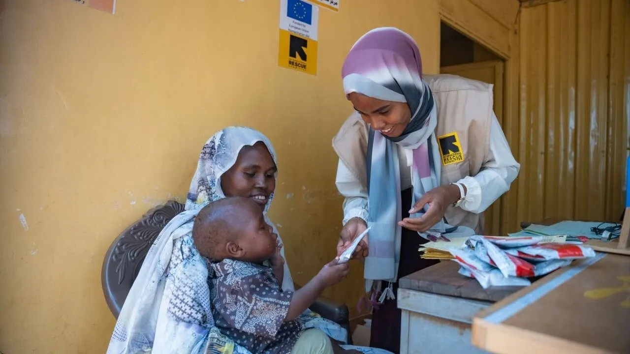 An IRC worker hands a nutrition supply to a young child sitting on an adult’s lap.
