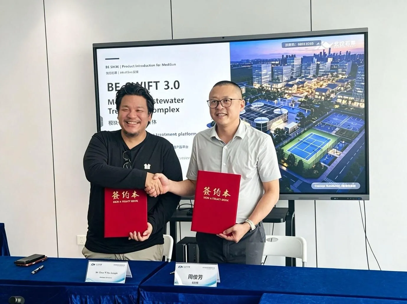 Two people shake hands while holding red contract folders in front of a presentation screen.