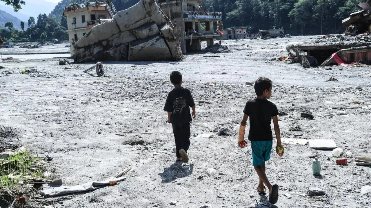 Two children walk through the debris-strewn site of their school in Nuwakot District, Nepal, with collapsed buildings behind them.