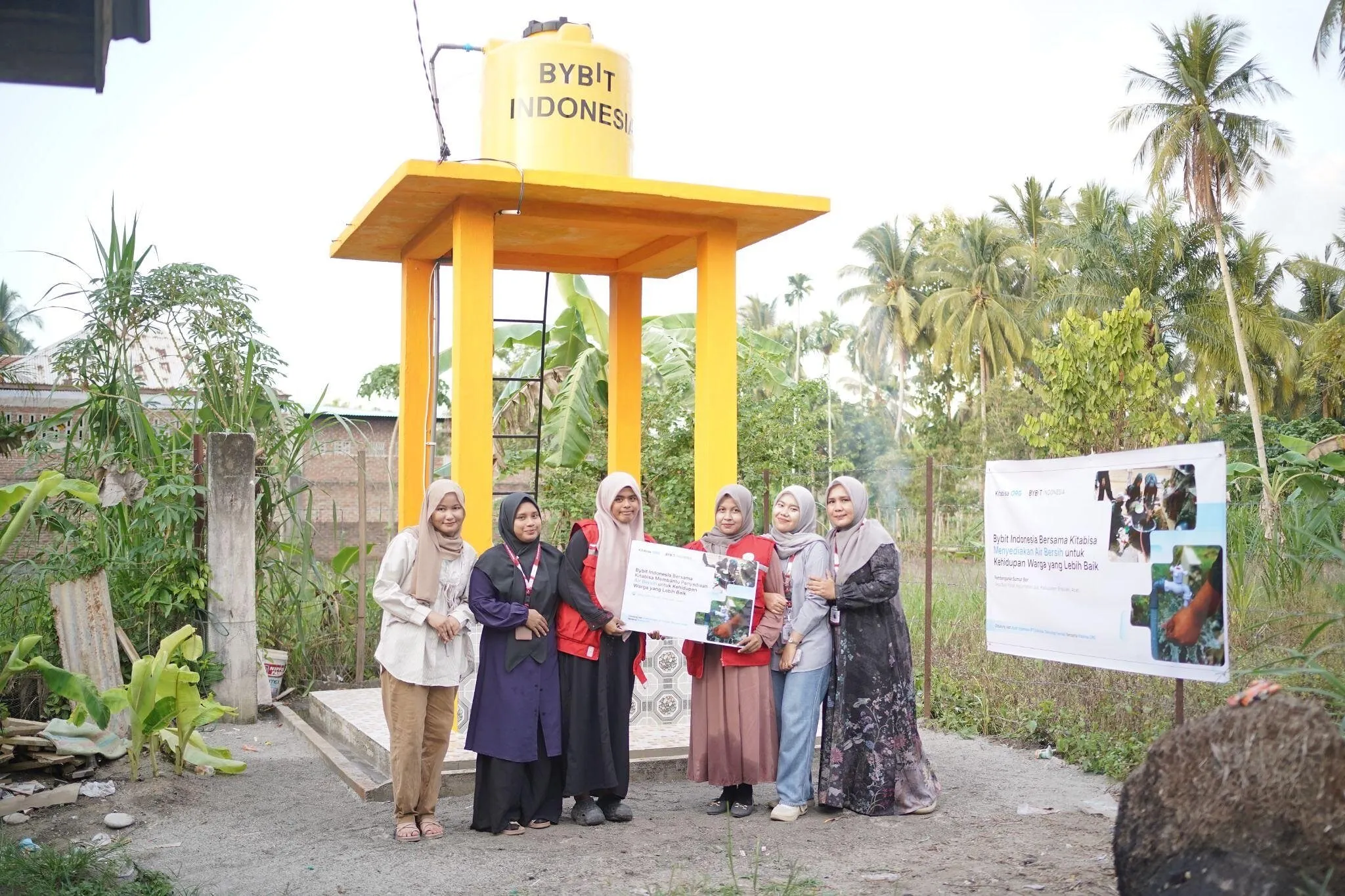 Six people stand beside a yellow water tower and a community water project sign.