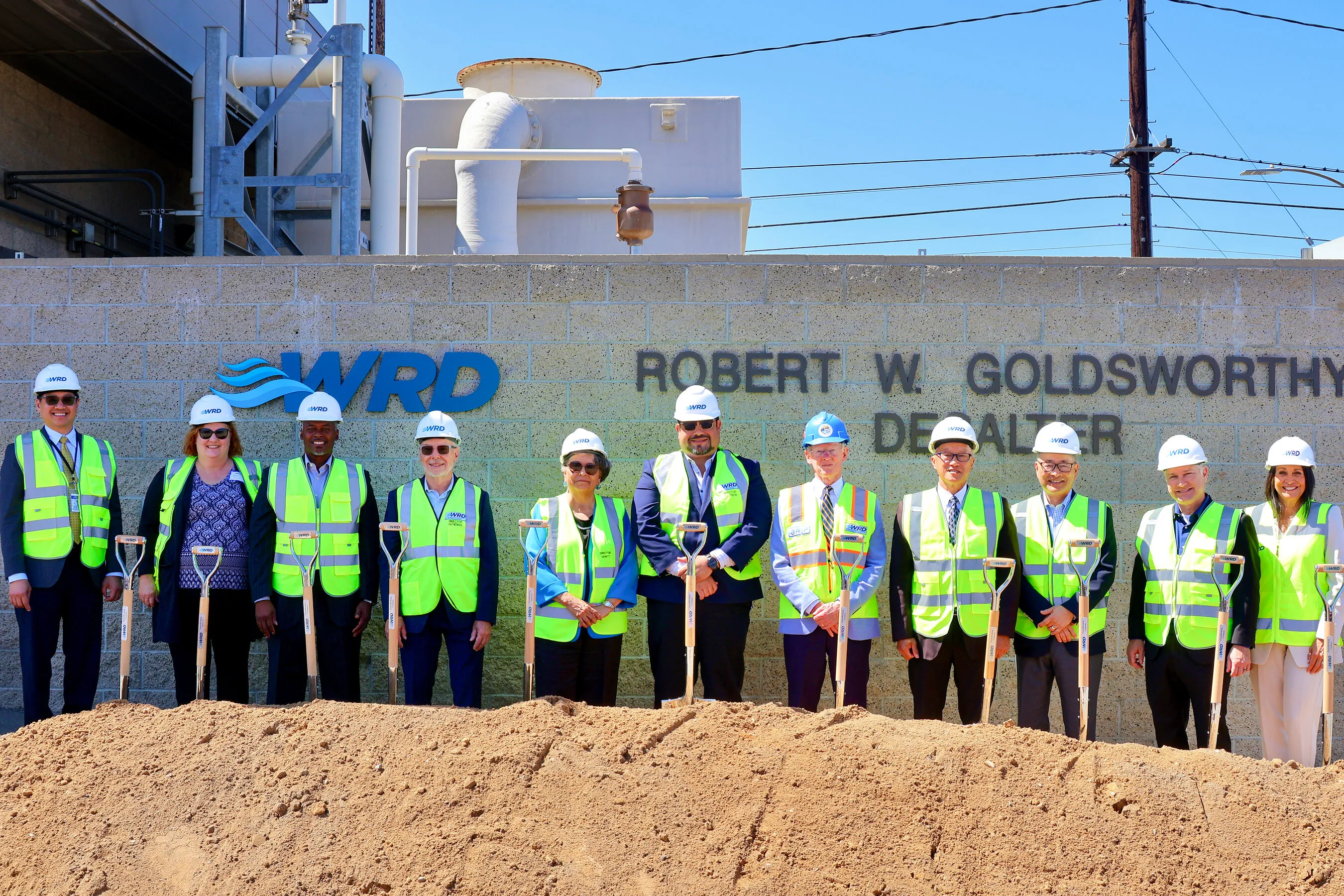 Eleven people in hard hats and safety vests hold shovels at the groundbreaking in front of the WRD Robert W. Goldsworthy Desalter sign.