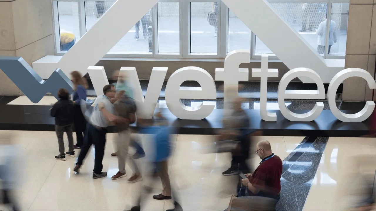 People walk past a large white WEFTEC sign inside a building.