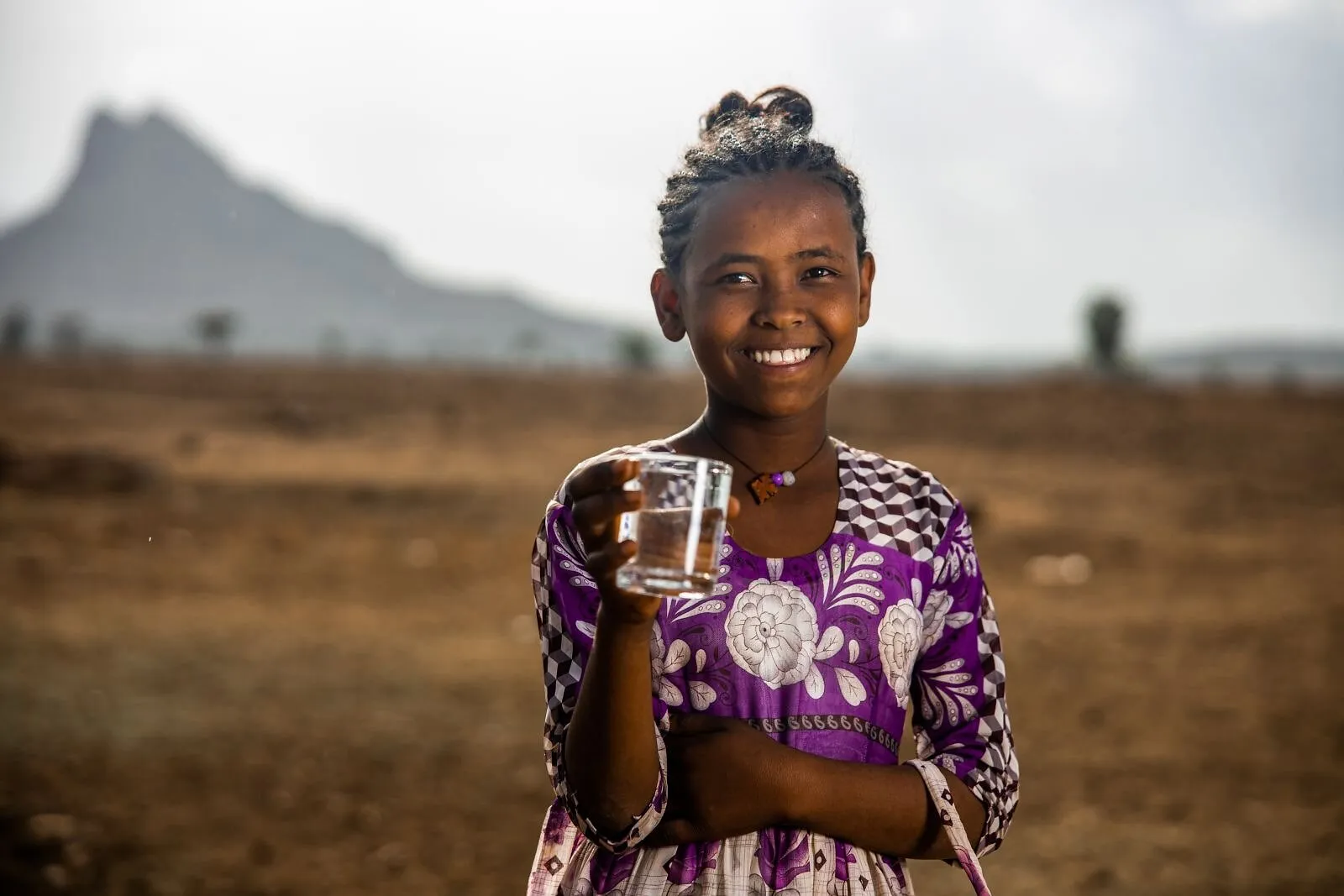 A smiling child holds a glass of water in a dry rural landscape.