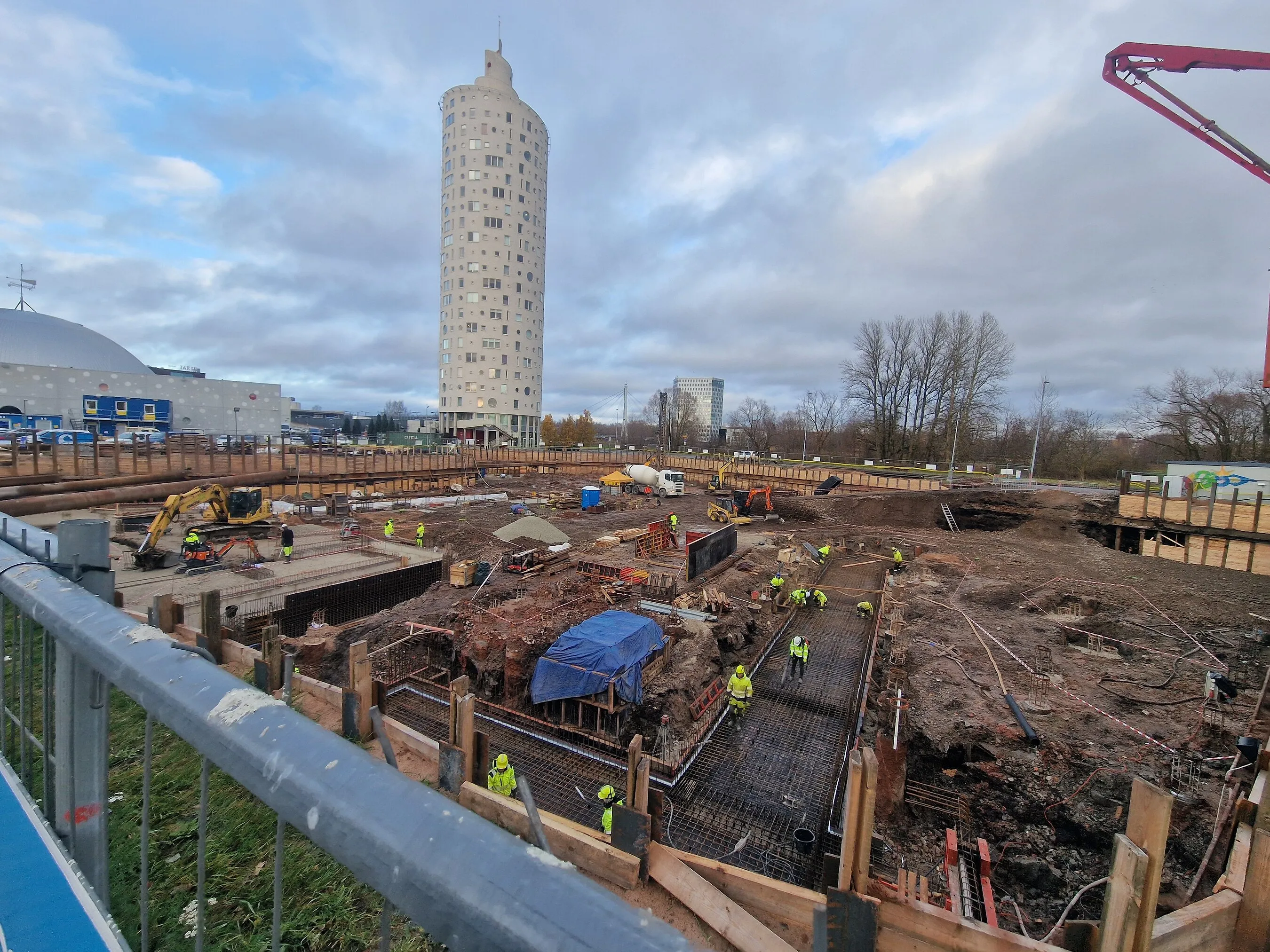 Construction workers excavate and reinforce foundations beside a tall cylindrical tower.