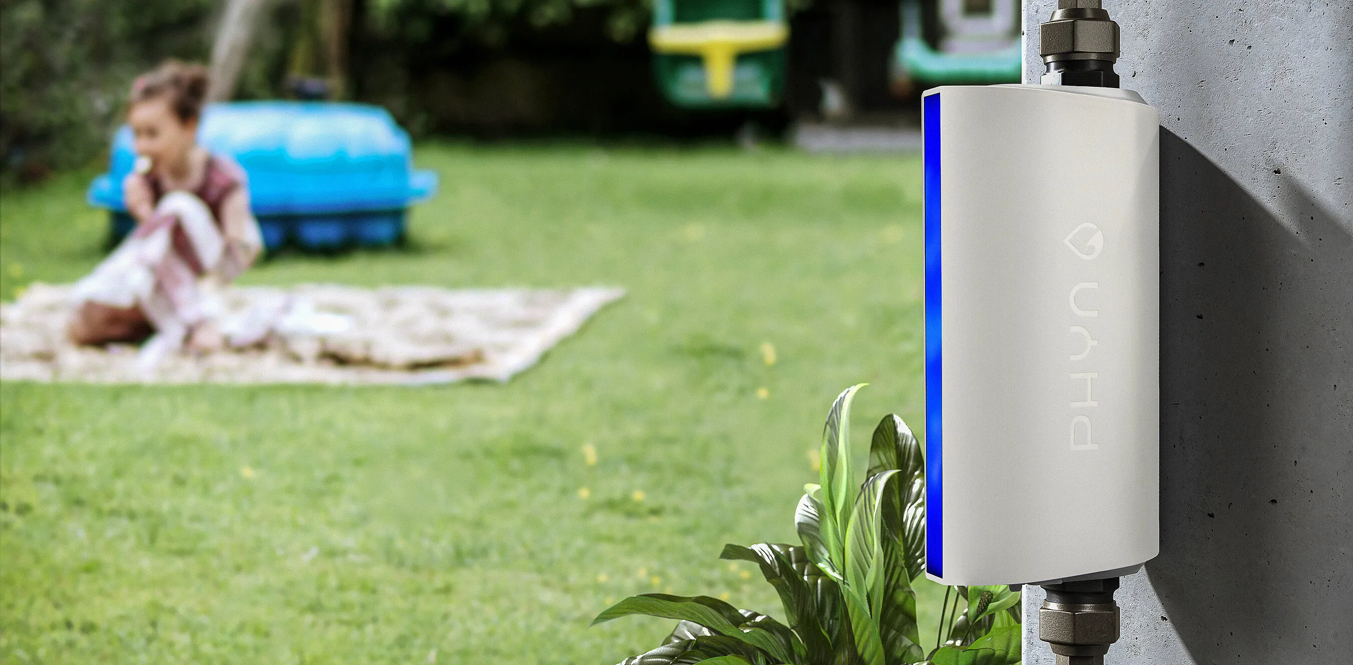 Phyn Protect installed on a pipe against a wall beside a lawn, with a child sitting on a blanket in the background