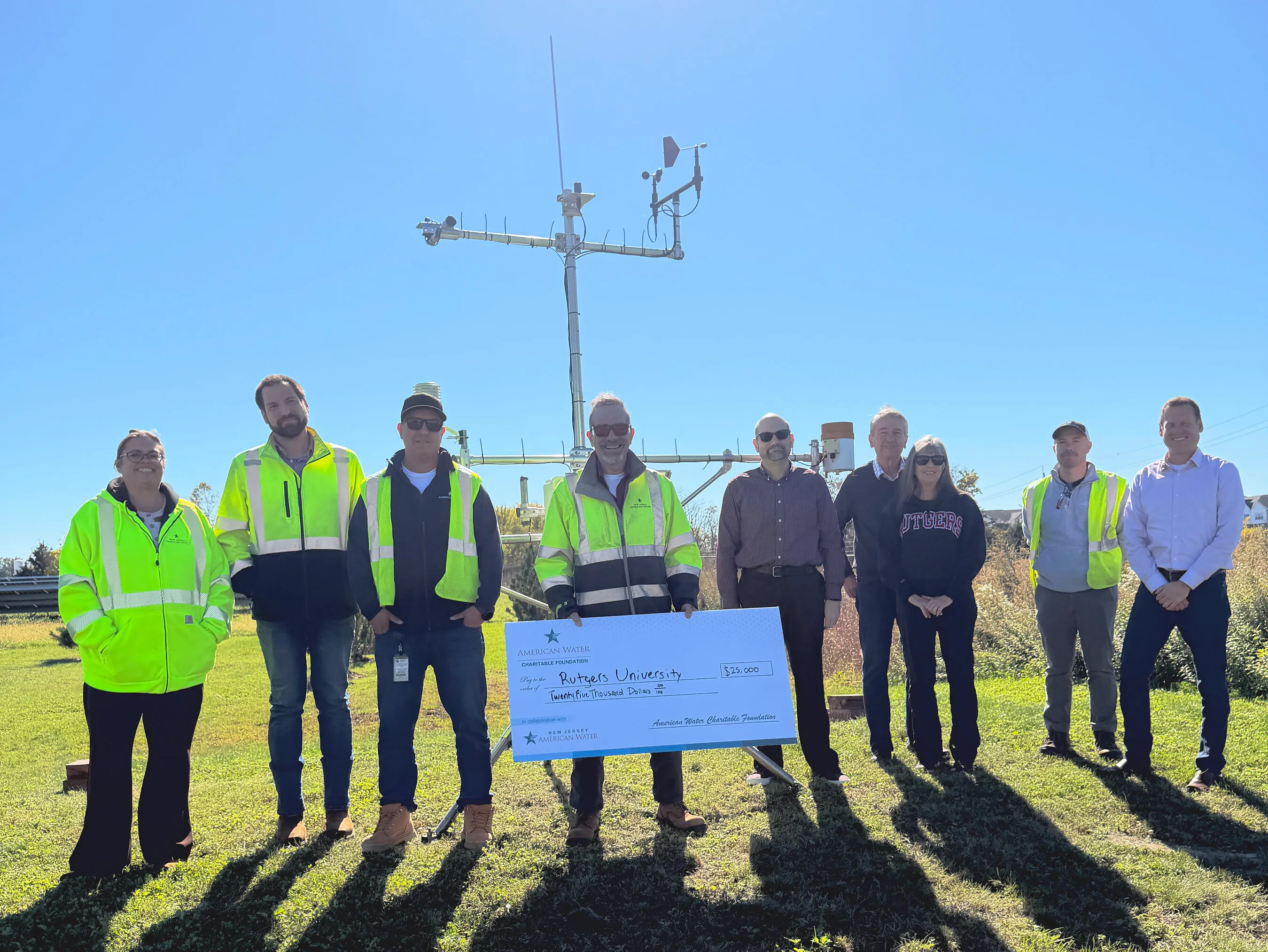 Nine people stand beside a Rutgers weather station, holding a ceremonial check.