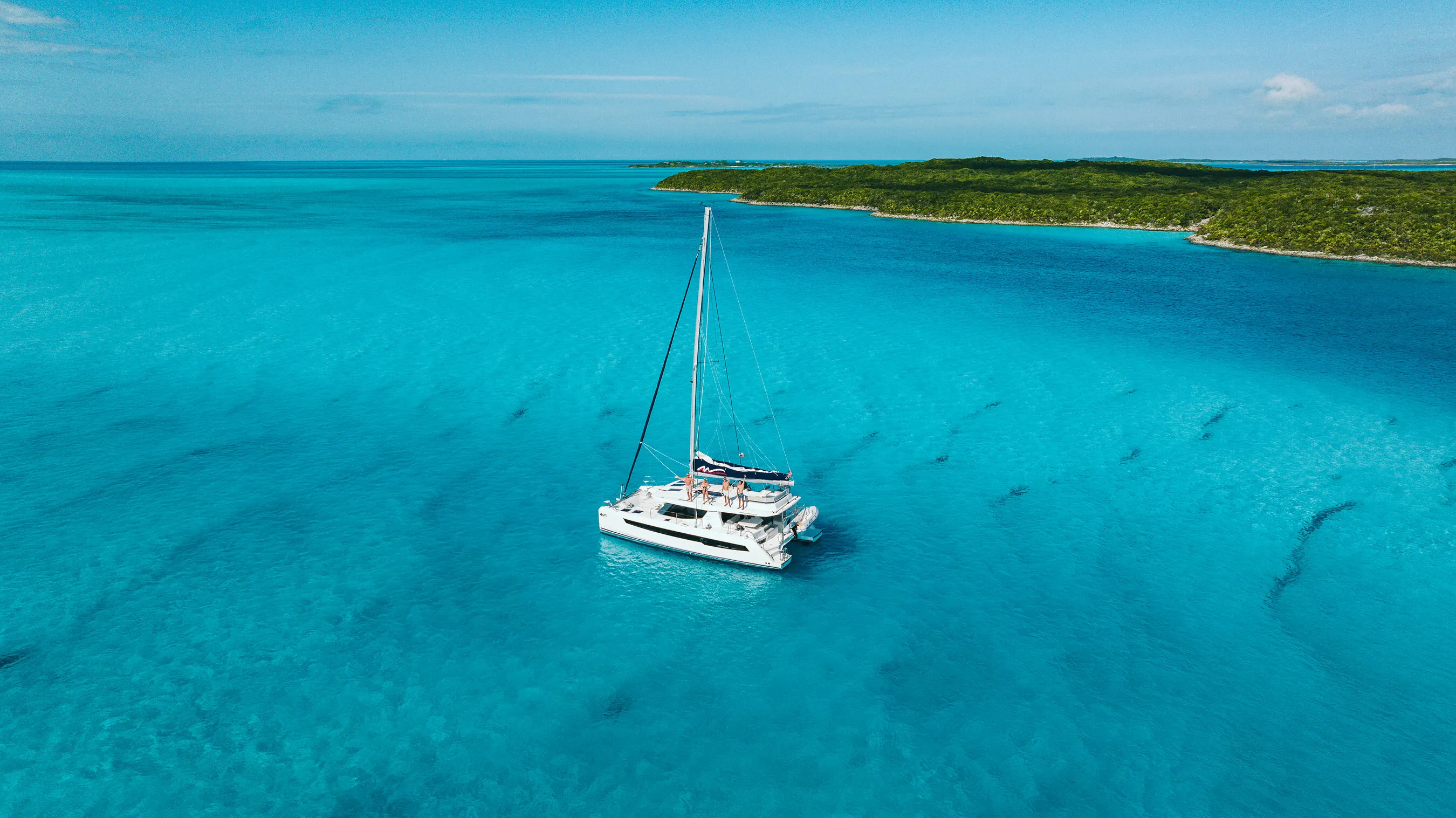 A catamaran sails on clear turquoise water beside a green island in the Bahamas.