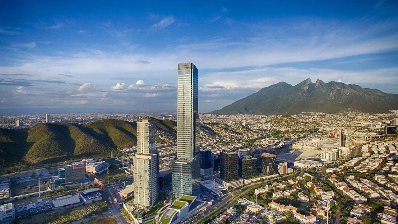 A tall tower rises above a dense cityscape, with mountains behind it.