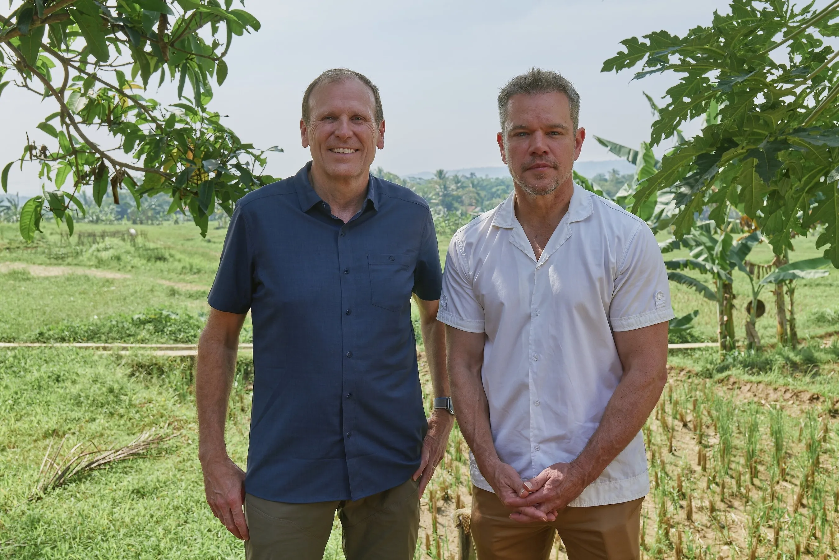 Gary White and Matt Damon standing in a green rural field beneath leafy trees.