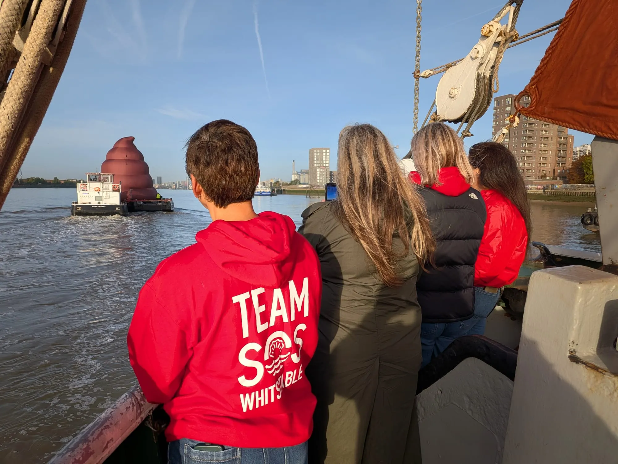 People in red and dark jackets face a brown inflatable sculpture on the river.