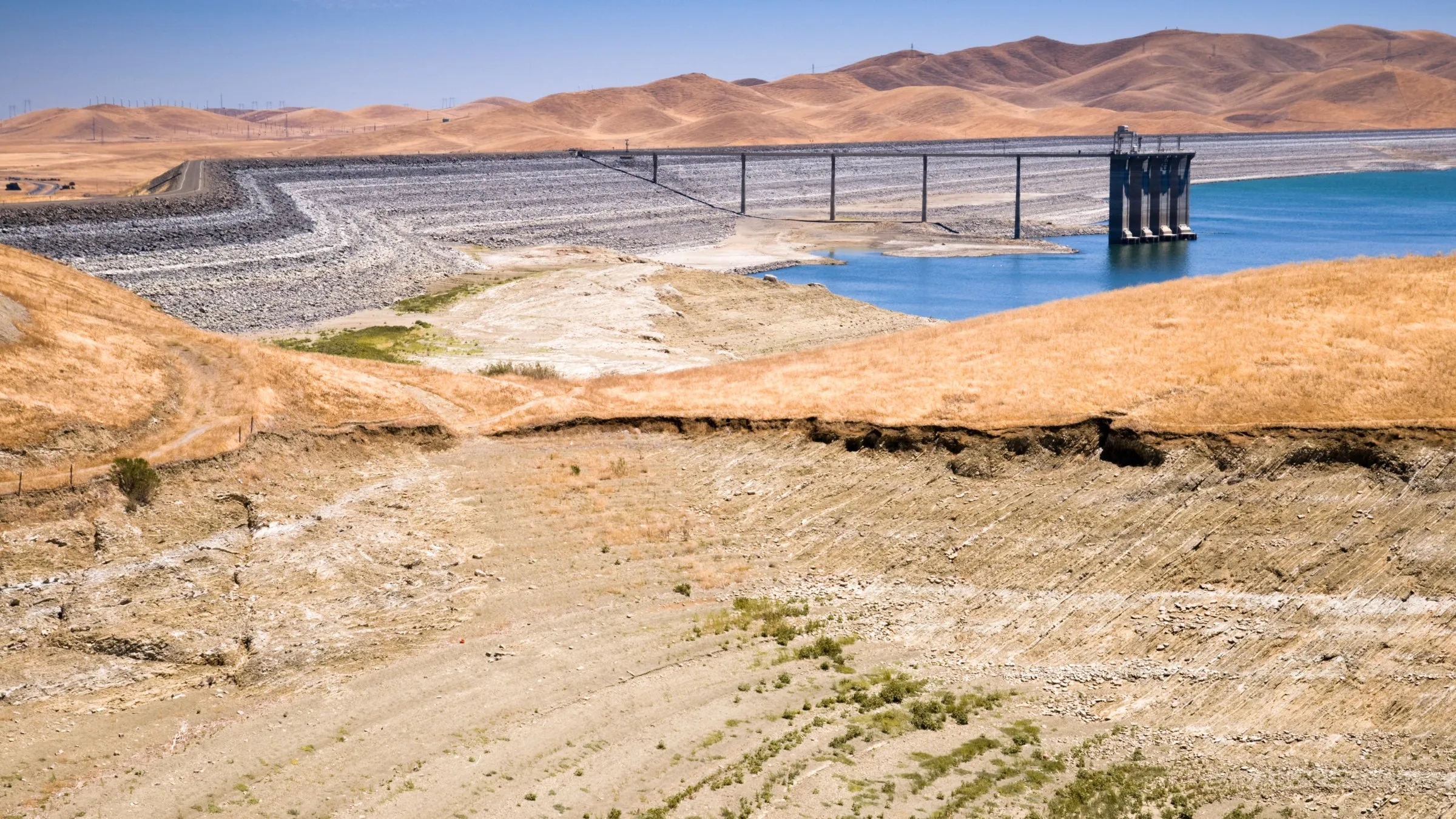 A reservoir with low water and dry, cracked banks below brown hills