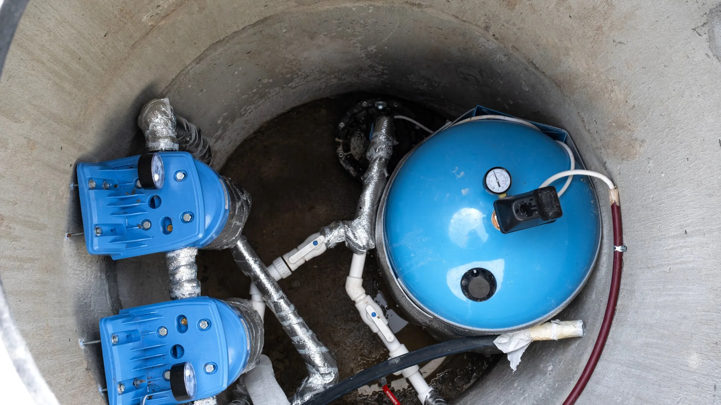 Looking down a well pit at blue pumps, a pressure tank and pipework