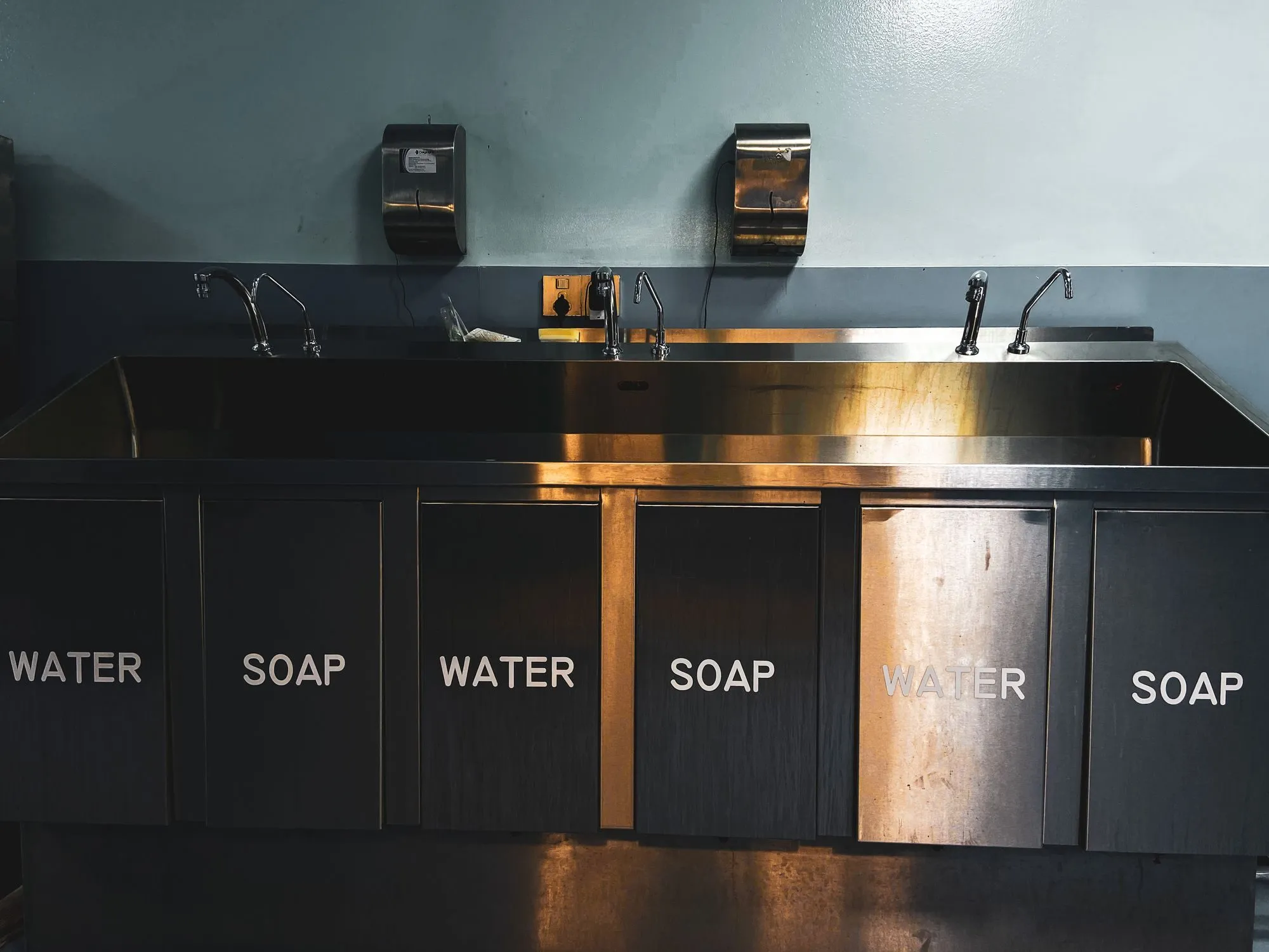 A row of steel hand-washing basins, each labelled water or soap
