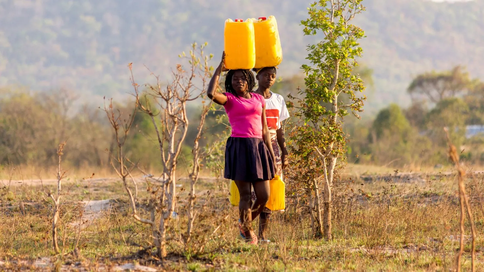 Two girls carrying yellow water containers on their heads across dry land