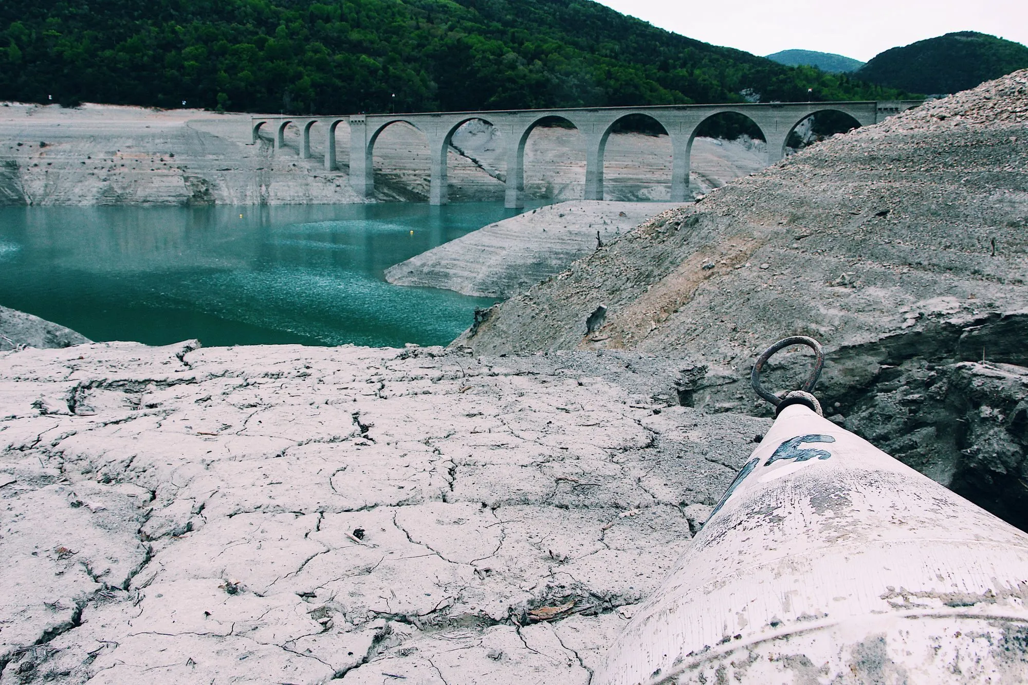 A dried-out reservoir bed, cracked and pale, beneath an old arched bridge