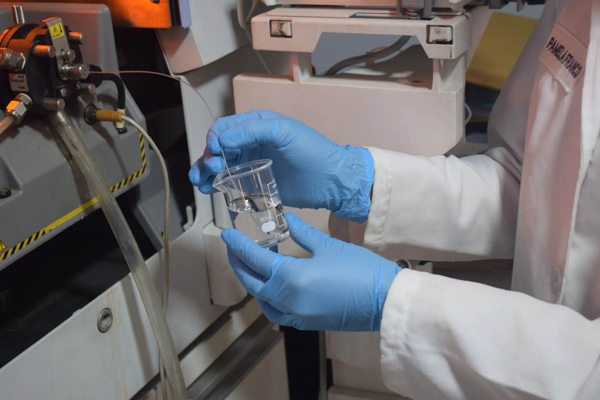 A laboratory worker in blue gloves holds a beaker of water beside testing equipment