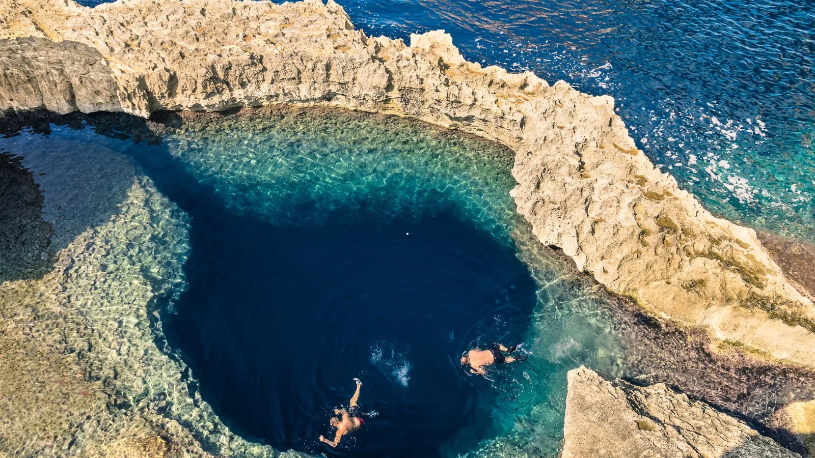 Swimmers in a deep blue sea pool among white rocks on the coast