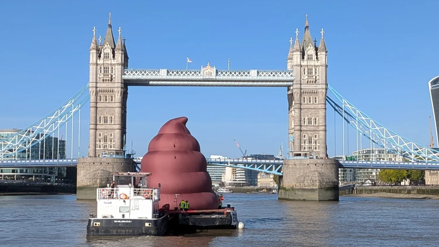 A huge brown inflatable sculpture stands on a barge in front of Tower Bridge.