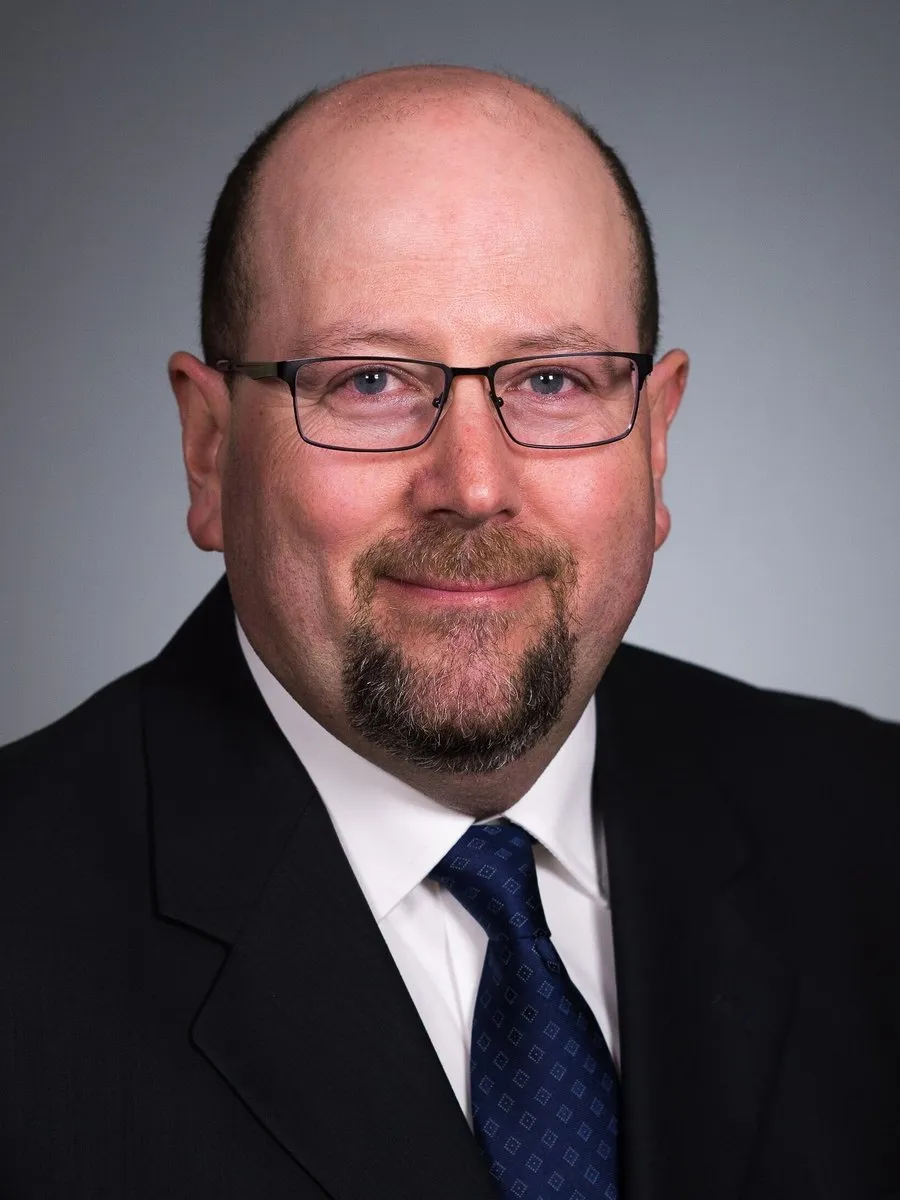 David Forcinito in glasses, a dark suit and blue tie against a gray background.
