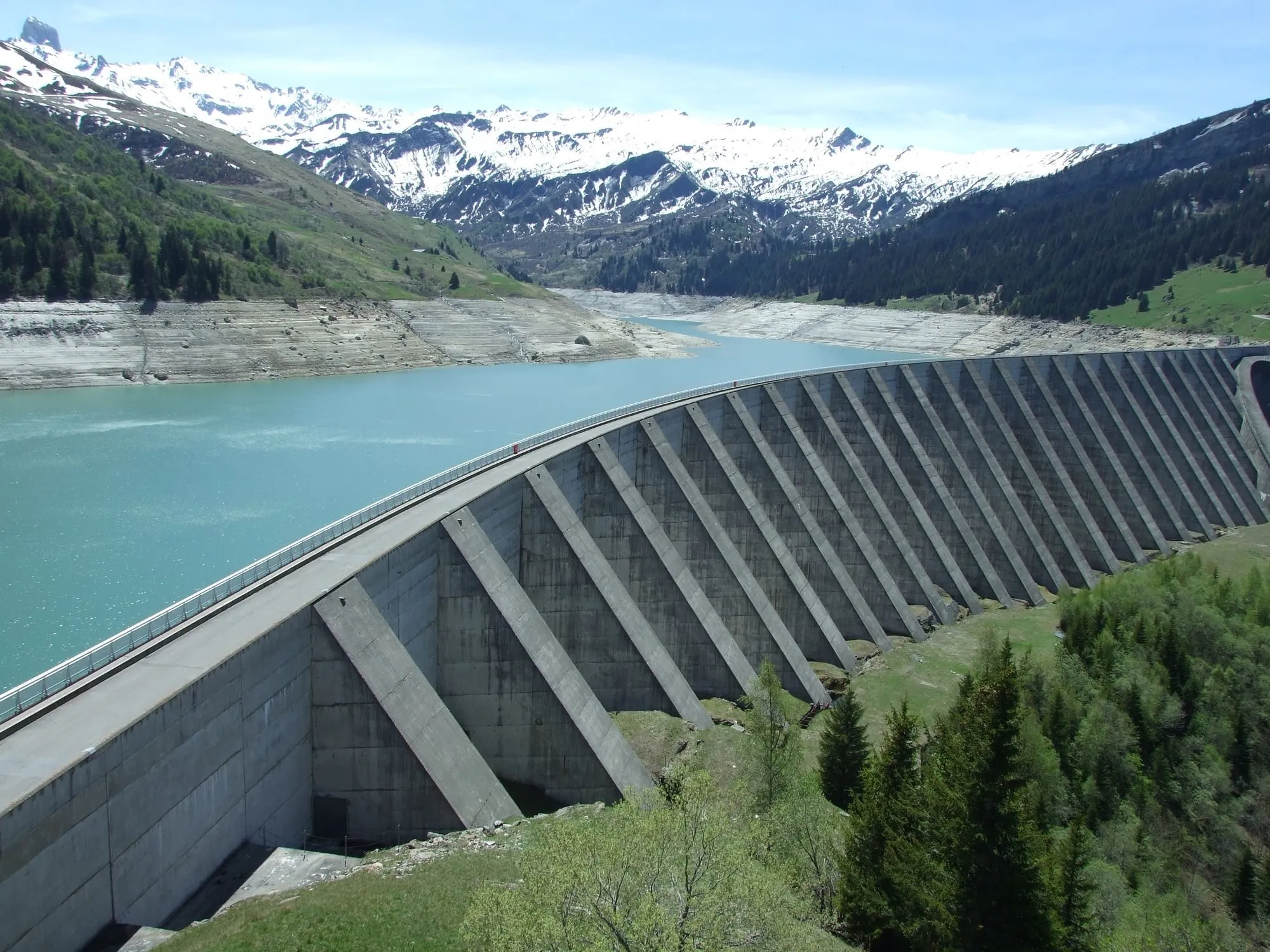 A concrete dam holding back a mountain reservoir under snow-capped peaks